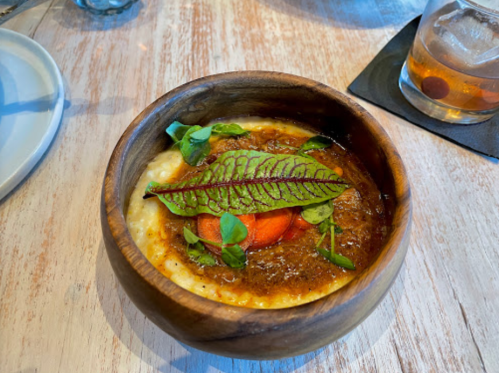 A wooden bowl filled with creamy dish topped with a leaf, herbs, and a slice of carrot, alongside a drink.
