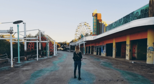 A person walks through an abandoned amusement park, with a Ferris wheel and colorful, graffiti-covered buildings in view.