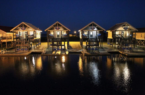 Four brightly lit cabins on stilts by a calm waterway at night, reflecting lights on the water's surface.