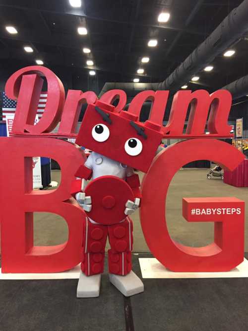 A red robot mascot stands between large letters spelling "Dream BIG" at an event, promoting positivity and creativity.