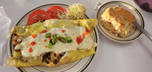 A plate with a large omelet filled with vegetables, topped with sauce, alongside sliced tomatoes and a piece of cornbread.
