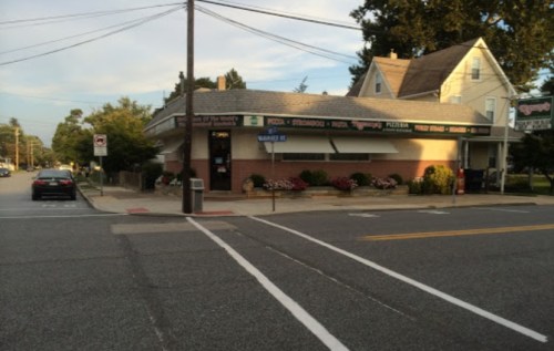 A corner restaurant with a sign for pizza and pasta, surrounded by flowers and trees, on a quiet street.