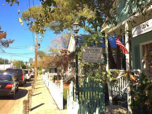 A sunny street scene featuring a shop sign for "History Antiques & Interiors" with trees and parked cars nearby.