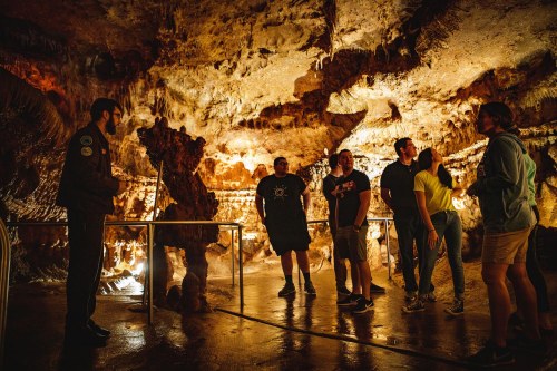 A tour guide speaks to a group of visitors in a dimly lit cave, surrounded by rock formations and illuminated by soft lights.