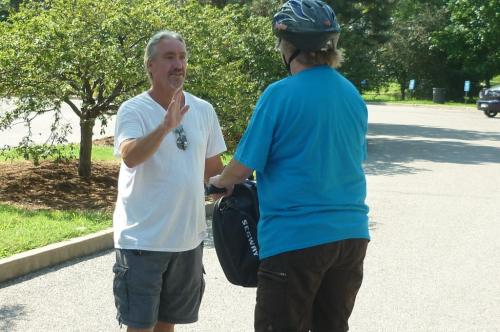 Two people converse outdoors; one is wearing a helmet and holding a bag, while the other gestures with a hand.