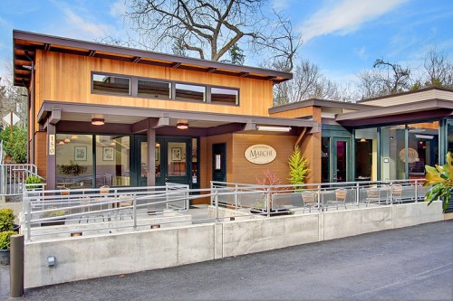 Modern wooden building with large windows, a sign reading "Marche," and a patio area surrounded by greenery.