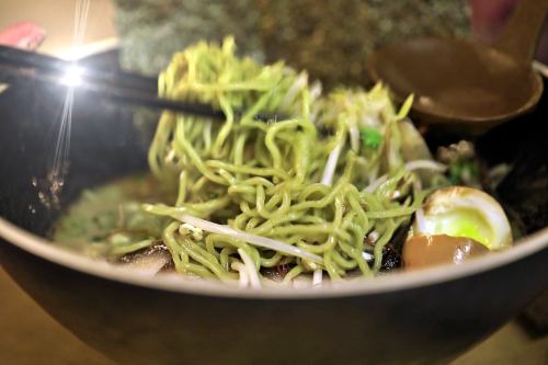 A close-up of a bowl of green noodles with vegetables and an egg, served with chopsticks.