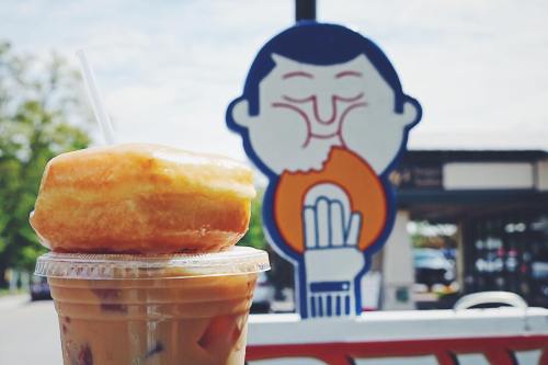 A donut on top of an iced coffee, with a cartoon sign of a person enjoying a donut in the background.