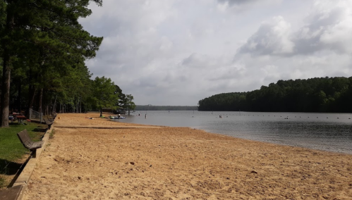 A sandy beach by a calm lake, surrounded by trees under a cloudy sky, with people enjoying the water in the distance.