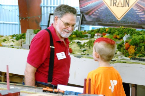 An older man with glasses and a beard talks to a young boy with red hair, surrounded by a model train display.