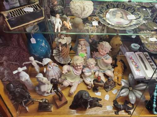 A display case filled with various antique dolls, figurines, and decorative items on a wooden shelf.