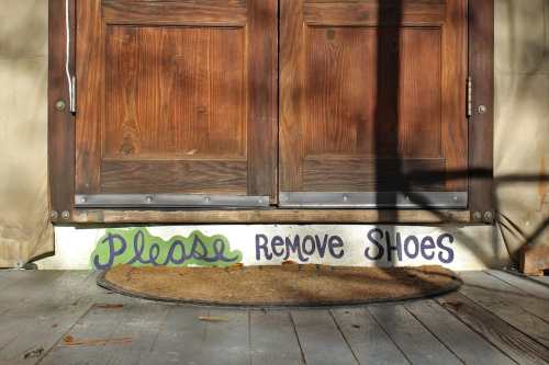 A wooden door with a sign that says "Please remove shoes" on the floor mat in front of it.