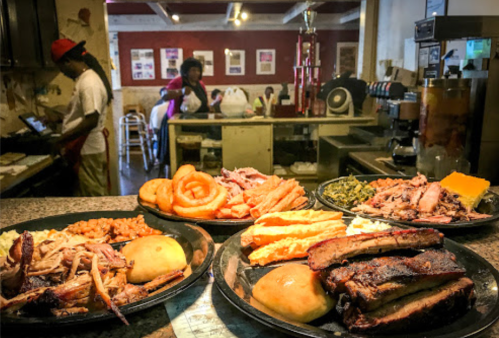A variety of barbecue dishes on plates, including ribs, pulled pork, sides, and a busy kitchen in the background.