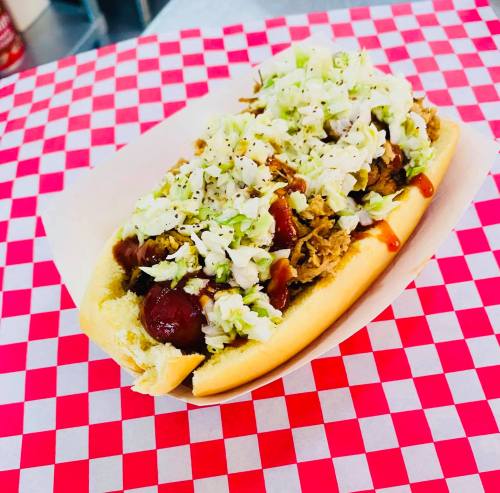 A hot dog topped with pulled pork and shredded lettuce, served in a bun on a red and white checkered background.