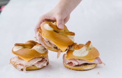 A hand reaching for a stack of three bagel sandwiches filled with turkey and cheese on a white surface.