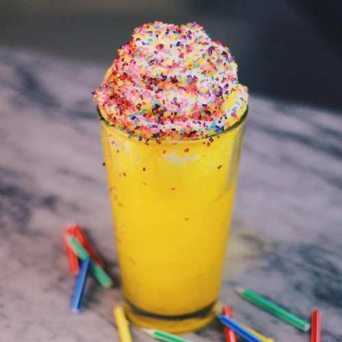 A vibrant yellow drink topped with whipped cream and colorful sprinkles, surrounded by small colorful straws.