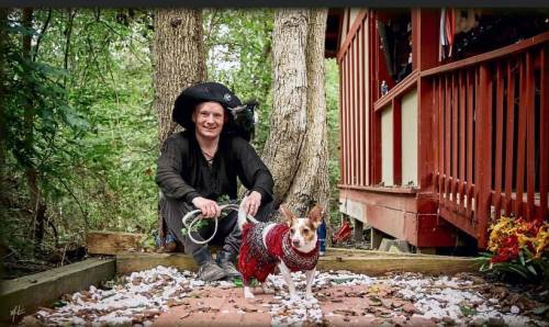 A person in a pirate costume kneels beside a small dog wearing a red outfit, surrounded by greenery and a wooden structure.