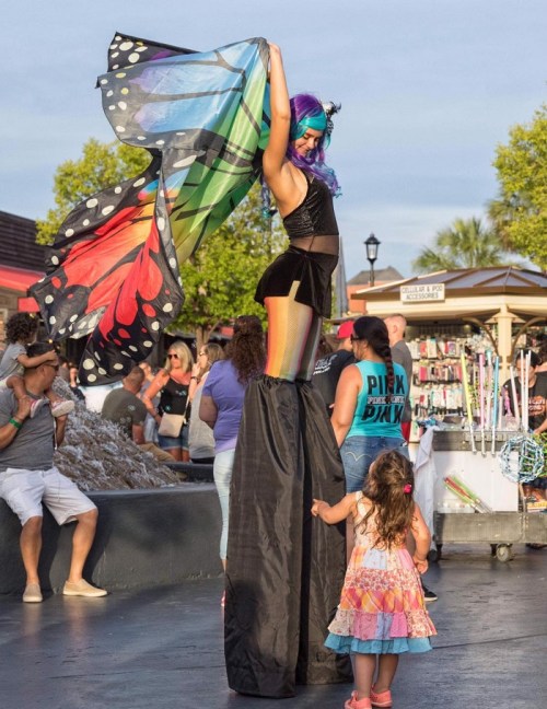 A performer on stilts with colorful hair and a butterfly cape interacts with a young girl at a lively outdoor event.