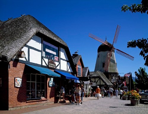 A charming street scene with thatched-roof buildings, a windmill, and people strolling under a clear blue sky.