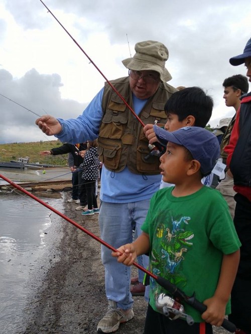 An adult teaches two children how to fish by a lake, with fishing rods in hand and a cloudy sky above.