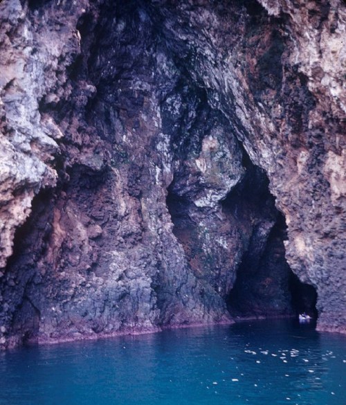 A rocky cave entrance by the sea, with calm blue water reflecting the surrounding cliffs.