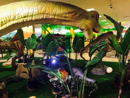 A colorful indoor scene featuring large dinosaur models among green plants and artificial grass.