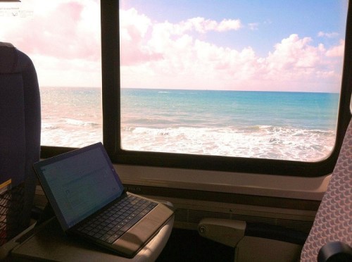 A laptop sits on a train seat, with a view of the ocean and blue sky through the window.