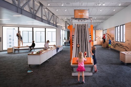 Children play in a bright, interactive space with a racing track, climbing wall, and various hands-on activities.