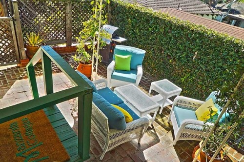 Cozy outdoor seating area with white wicker furniture, colorful cushions, and greenery in a sunny garden setting.