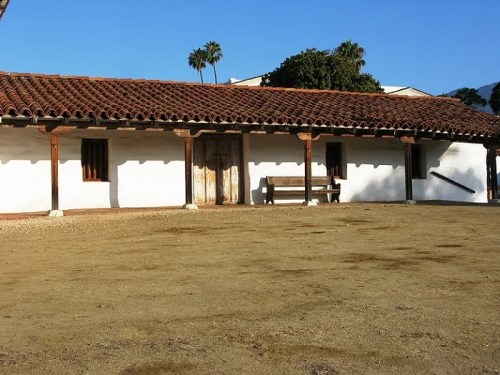 A historic building with a tiled roof, white walls, and a dirt courtyard, surrounded by palm trees under a clear blue sky.