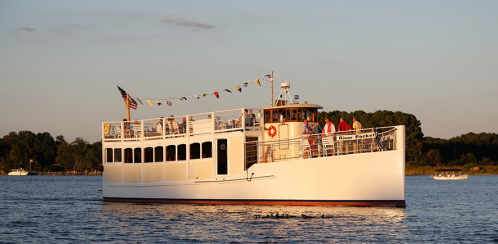 A white boat with passengers sails on calm water, surrounded by trees and a clear sky at sunset.