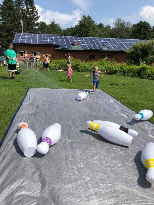 A child runs on a water slide with inflatable bowling pins, while others play in the background near a solar-paneled building.