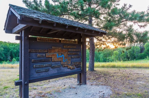 Sign at Hayes Lake State Park displaying park information and a map, with trees and a sunset in the background.