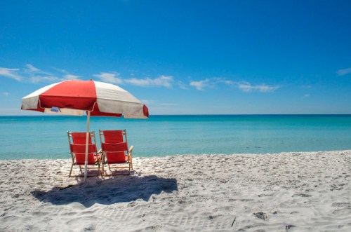 Two red and white beach chairs under an umbrella on a sandy beach, with calm turquoise waters and a clear blue sky.