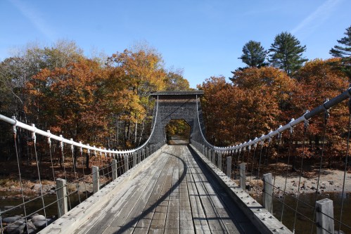 A wooden suspension bridge leads through colorful autumn trees under a clear blue sky.
