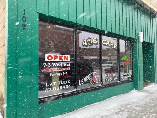 A green building with a cafe window displaying hours, location, and "OPEN" signs, amidst falling snow.