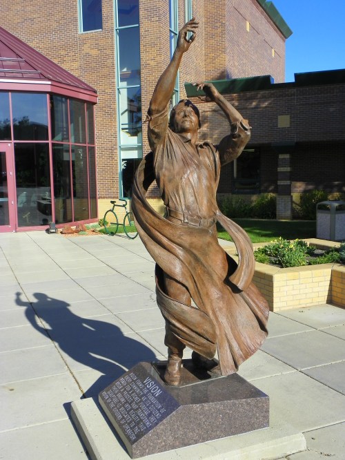A bronze statue of a figure with outstretched arms, draped in flowing fabric, stands outside a building.