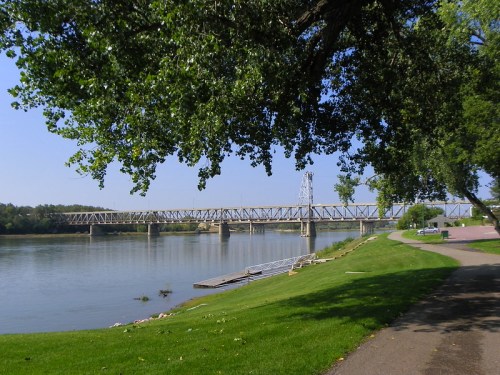 A scenic view of a river with a bridge in the background, surrounded by green trees and a walking path.