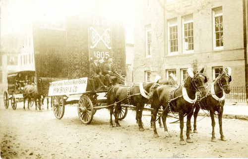 A vintage horse-drawn delivery wagon with two horses, parked on a street, featuring the year 1905 and company name.