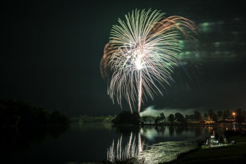 Colorful fireworks burst over a calm lake at night, reflecting light on the water's surface. Trees and lights are visible nearby.