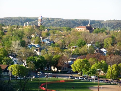 A scenic view of a small town with green trees, houses, and a distant clock tower on a sunny day.