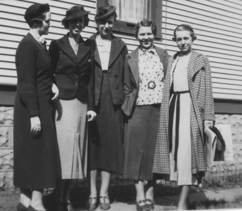 Five women in vintage clothing and hats stand together outside a house, smiling at the camera.