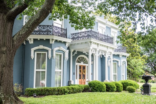 A stately blue house with white trim, balconies, and a lush green lawn, framed by trees and decorative landscaping.