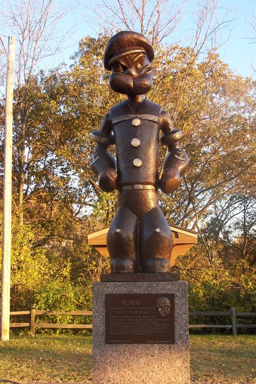 A bronze statue of Popeye the Sailor, standing proudly on a stone pedestal, surrounded by trees.