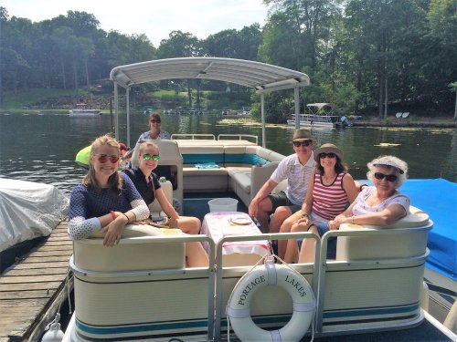 A group of six people enjoying a sunny day on a boat, surrounded by trees and water, with snacks on the table.