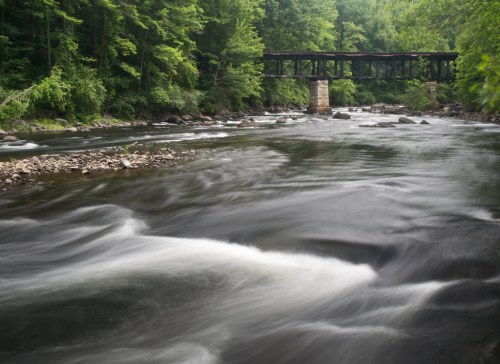 A serene river flows beneath a rustic bridge, surrounded by lush green trees and rocky banks.