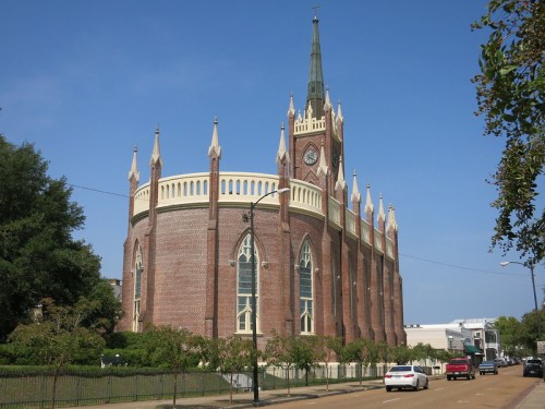 A large brick church with a tall spire and clock, surrounded by trees and a clear blue sky.