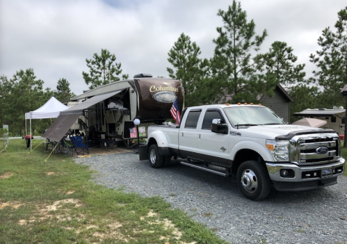 A white truck parked next to a large RV in a grassy campsite, surrounded by trees and a cloudy sky.