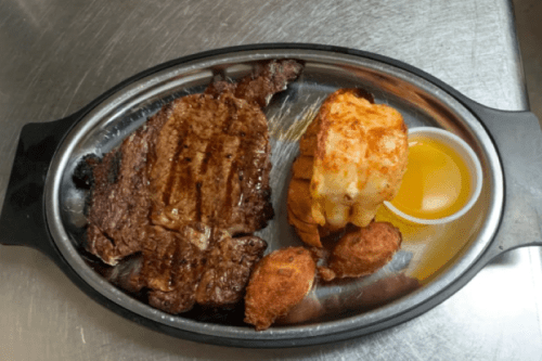 A sizzling steak on a platter with a side of shrimp and dipping sauce, accompanied by two fried items.