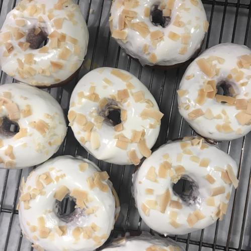A close-up of freshly baked donuts topped with white icing and coconut flakes, arranged on a cooling rack.